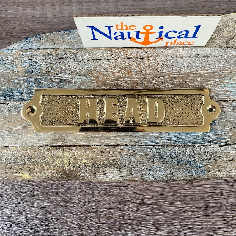 May include: A gold-coloured brass sign with the word "HEAD" embossed on it. The sign has a textured surface and rounded edges. It is mounted on a wooden surface.