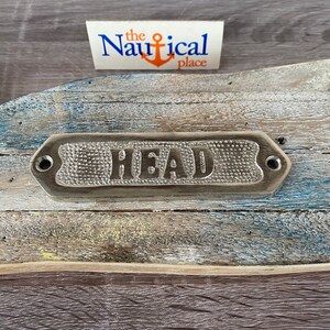 May include: A silver metal sign with the word "HEAD" embossed on it. The sign is mounted on a weathered wooden surface.