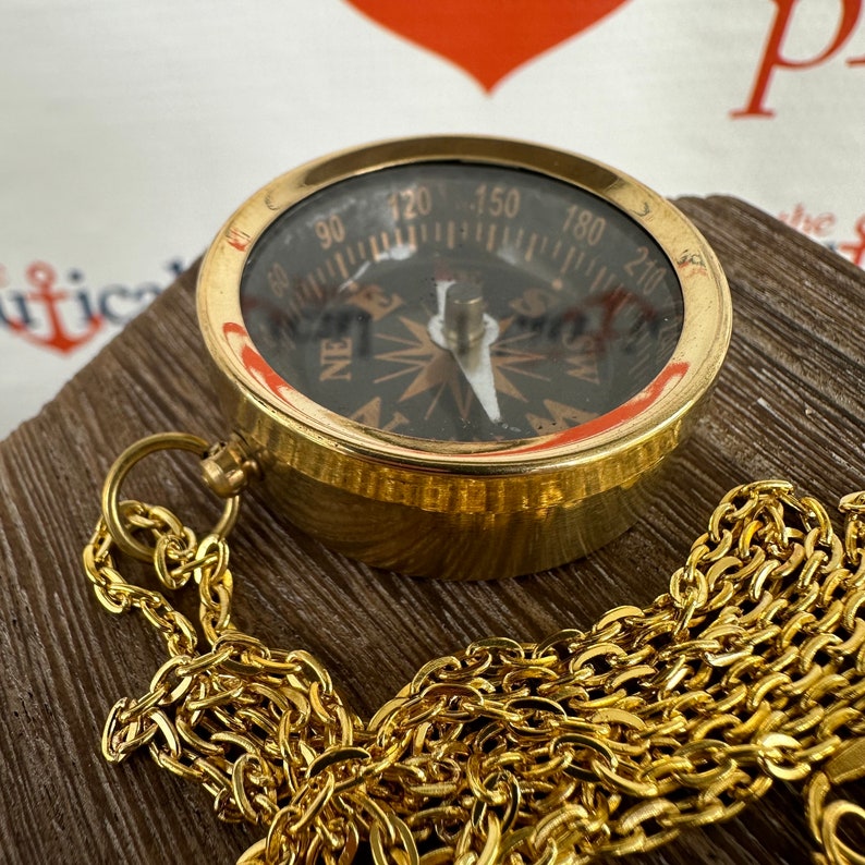 May include: A brass compass with a black face and a gold chain. The compass has a needle that points to north, south, east, and west. The chain is attached to the compass with a loop.