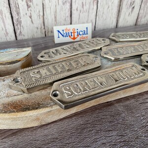 May include: A set of silver metal nautical door signs with various room names, including "Captain", "Ship's" and "Shower Room". The signs are on a wooden surface with a small sign that reads "the Nautical place".