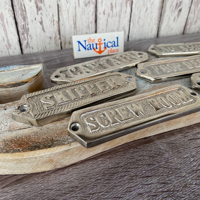 May include: A set of silver metal nautical door signs with various room names, including "Captain", "Ship's" and "Shower Room". The signs are on a wooden surface with a small sign that reads "the Nautical place".