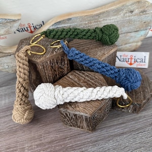 May include: Four different colored rope keychains with a knot at the end. The keychains are brown, white, green, and blue. The keychains are displayed on a wooden surface. The text "the Nautical place" is visible in the background.