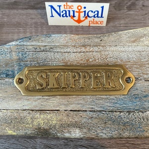 May include: Brass plaque with the word "SKIPPER" embossed on it. The plaque has rounded edges and two small holes on either side.