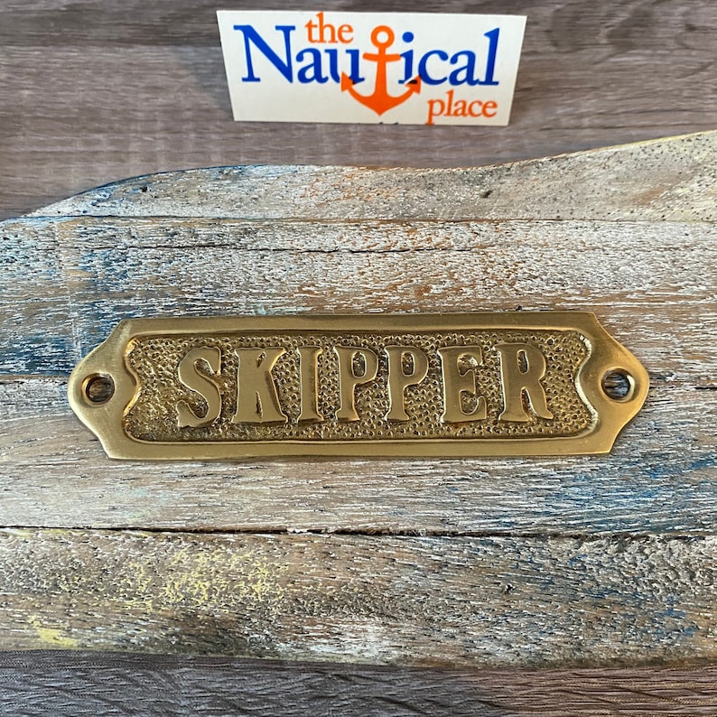 May include: Brass plaque with the word "SKIPPER" embossed on it. The plaque has rounded edges and two small holes on either side.