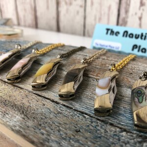 Pu&ograve; includere: Sei diversi ciondoli a forma di coltellino da tasca intarsiati con conchiglie su catene dorate. I ciondoli sono tutti di colori e motivi diversi. Il testo "The Nautical Place" &egrave; visibile sullo sfondo.