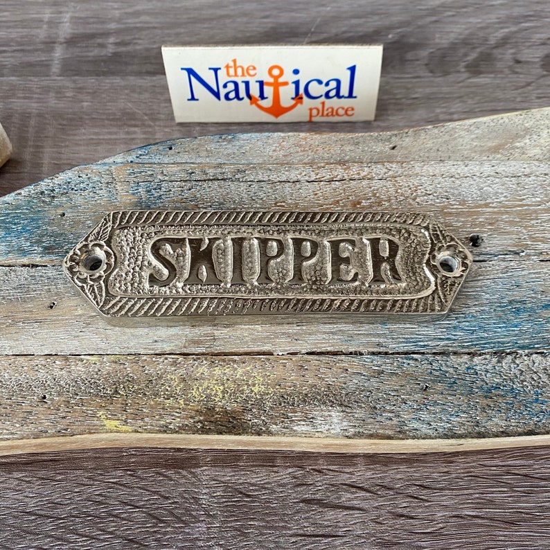 May include: A silver metal sign with the word "SKIPPER" embossed on it. The sign is attached to a weathered wooden surface.