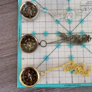 May include: Three gold compass necklaces with different chain styles. The compasses are all round and have a black face with white markings. The chains are silver, bronze, and gold.