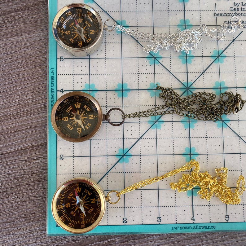 May include: Three gold compass necklaces with different chain styles. The compasses are all round and have a black face with white markings. The chains are silver, bronze, and gold.