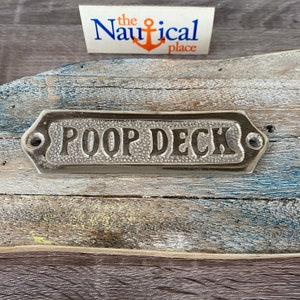 May include: A silver metal sign with the words "Poop Deck" embossed on it. The sign is mounted on a weathered wooden surface.