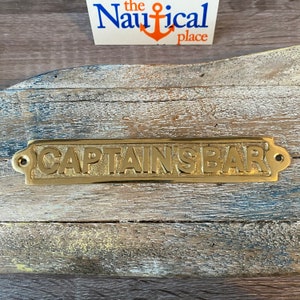 May include: Brass sign with the words "CAPTAIN'S BAR" in raised lettering. The sign has rounded corners and two small holes on either side.