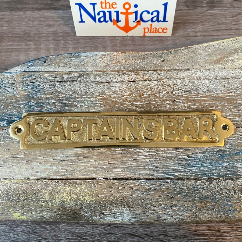 May include: Brass sign with the words "CAPTAIN'S BAR" in raised lettering. The sign has rounded corners and two small holes on either side.