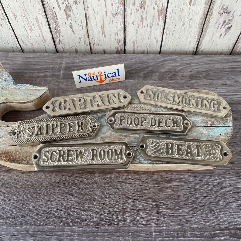 May include: Seven decorative metal signs with nautical themed words. The signs are in a variety of shapes and sizes and have a brushed silver finish. The words on the signs are "Captain", "Skipper", "Screw Room", "Poop Deck", "Head", "No Smoking", and "The Nautical Place".