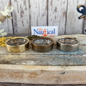 May include: Three round compasses with black faces and gold numbers. The compasses are in brass, bronze, and silver finishes. The compasses are on a wooden surface.