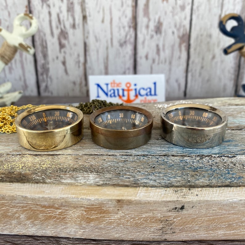 May include: Three round compasses with black faces and gold numbers. The compasses are in brass, bronze, and silver finishes. The compasses are on a wooden surface.