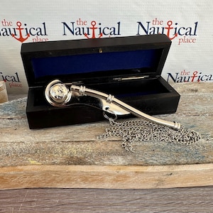 Puede incluir: Un silbato de barco plateado con un ancla en la parte superior, en una caja de madera negra con forro de terciopelo azul. El silbato está unido a una cadena plateada.