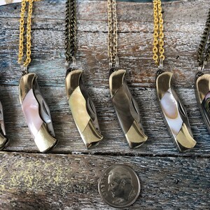 Pu&ograve; includere: Sei coltelli tascabili argentati con intarsi in madreperla di diversi colori sono esposti su una superficie di legno. Ogni coltello &egrave; attaccato a una collana a catena dorata.