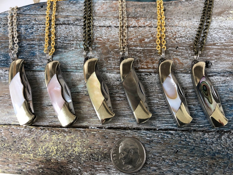 Pu&ograve; includere: Sei coltelli tascabili argentati con intarsi in madreperla di diversi colori sono esposti su una superficie di legno. Ogni coltello &egrave; attaccato a una collana a catena dorata.