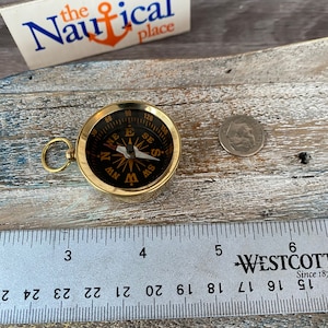 May include: A small, gold-coloured compass with a black face and a white needle. The compass is approximately 5 cm in diameter and has a ring around the edge with numbers from 0 to 180. The compass is resting on a wooden surface.