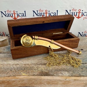 Puede incluir: Un silbato náutico de color dorado con un diseño de ancla en la boquilla. El silbato está unido a una cadena dorada y se encuentra en una caja de madera con el texto "the Nautical place" en la tapa.
