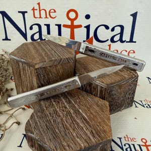 May include: Two silver crab knives with etched designs and wooden handles are displayed on wooden blocks. One knife reads "CARVEL HALL" and the other reads "CRAB KNIFE". The background features nautical-themed text and imagery.