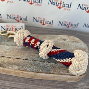 May include: A red, white, and blue rope keychain with a knot at the end. The keychain is made of natural rope and has a gold metal ring.