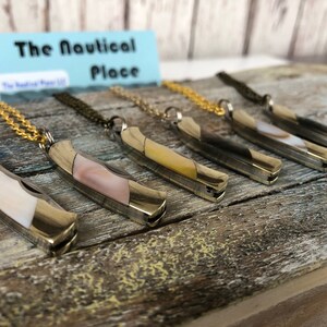 Pu&ograve; includere: Sei coltelli tascabili argentati con intarsi in madreperla di diversi colori sono esposti su una superficie di legno. Ogni coltello &egrave; attaccato a una collana a catena dorata. Il cartello sopra i coltelli recita "The Nautical Place".