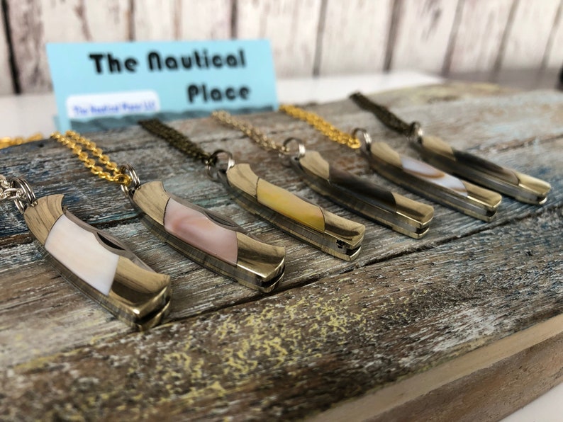 Pu&ograve; includere: Sei coltelli tascabili argentati con intarsi in madreperla di diversi colori sono esposti su una superficie di legno. Ogni coltello &egrave; attaccato a una collana a catena dorata. Il cartello sopra i coltelli recita "The Nautical Place".
