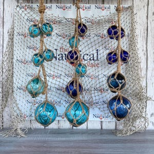 May include: Three vertical strands of decorative glass fishing floats in shades of blue and turquoise, each encased in tan jute netting. The floats are suspended against a background featuring a white fishing net and the text the Nautical place.