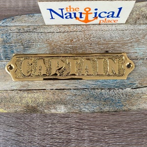 May include: A gold-coloured metal sign with the word "CAPTAIN" embossed on it. The sign has a slightly textured surface and rounded edges.