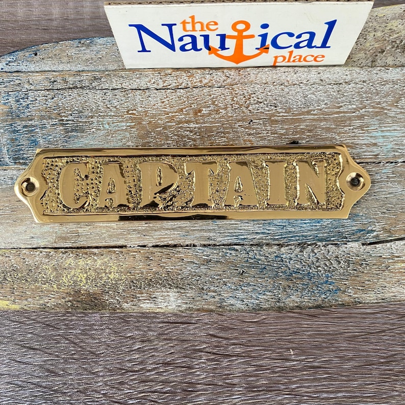 May include: A gold-coloured metal sign with the word "CAPTAIN" embossed on it. The sign has a slightly textured surface and rounded edges.