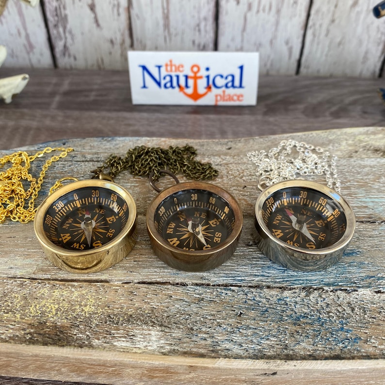 May include: Three antique-style compass necklaces with gold, brass, and silver chains. Each compass has a black face with gold markings and a red needle. The compasses are on a wooden surface with the text "the Nautical place" on a card in the background.