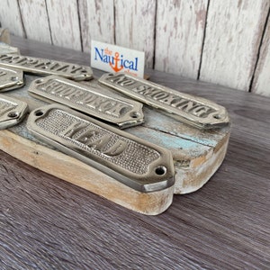 May include: A set of six silver-colored metal signs with nautical themed words. The signs are arranged on a weathered wooden surface. The words on the signs are "Head", "Galleon", "Captain", "No Smoking", "Forward", and "Aft".