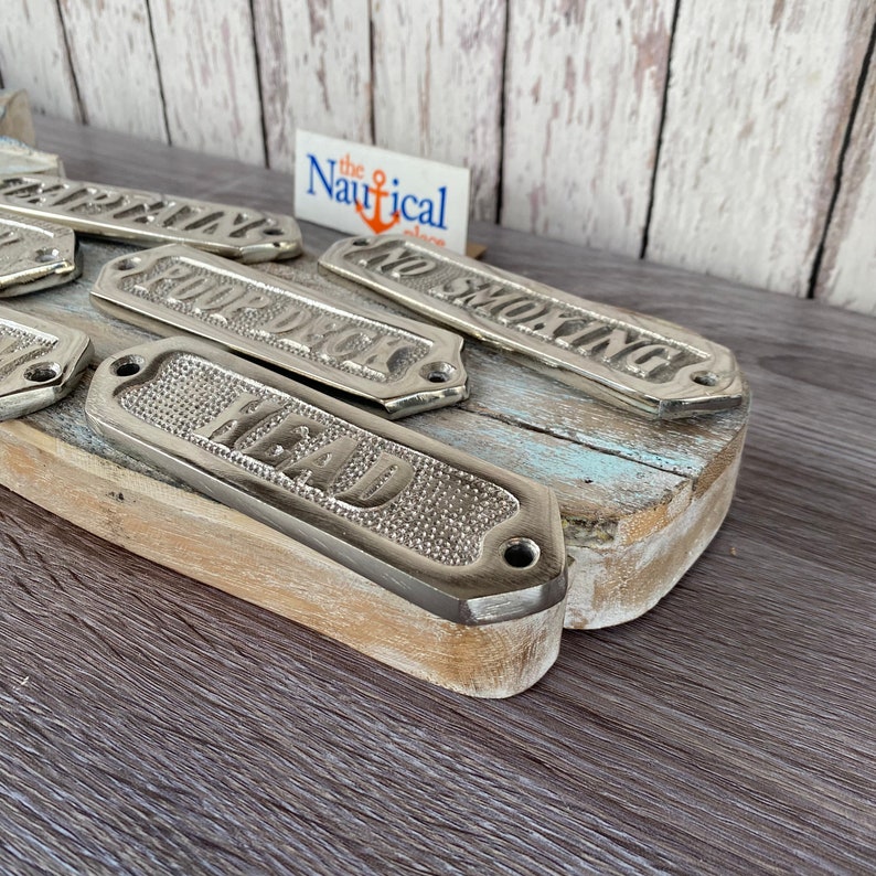 May include: A set of six silver-colored metal signs with nautical themed words. The signs are arranged on a weathered wooden surface. The words on the signs are "Head", "Galleon", "Captain", "No Smoking", "Forward", and "Aft".