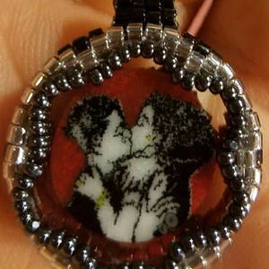 May include: A handmade pendant featuring a black and white image of two people kissing, set within a beaded frame. The frame is made of black and silver beads, with a red background. The pendant has a loop at the top for a chain.