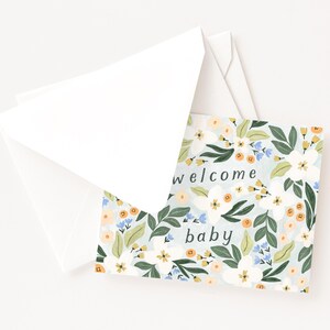 Op de afbeelding: Een wenskaart met een bloemenontwerp en de woorden "welcome baby" erop gedrukt. De kaart heeft een lichtblauwe achtergrond met witte, oranje en blauwe bloemen en groene bladeren. Het wordt vergezeld door een witte envelop.