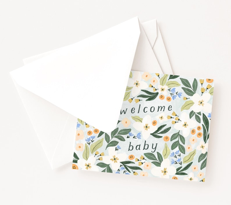 Op de afbeelding: Een wenskaart met een bloemenontwerp en de woorden "welcome baby" erop gedrukt. De kaart heeft een lichtblauwe achtergrond met witte, oranje en blauwe bloemen en groene bladeren. Het wordt vergezeld door een witte envelop.