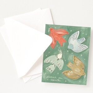 Pu&ograve; includere: Un biglietto di auguri con quattro colombe illustrate in rosso, blu e oro su sfondo verde. Il biglietto recita "peace on earth". Buste bianche sono visibili accanto al biglietto.
