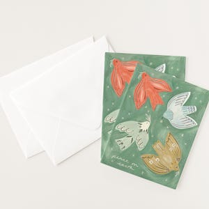 Pu&ograve; includere: Biglietti di auguri con illustrazioni di colombe rosse, blu e oro su sfondo verde. I biglietti sono accompagnati da buste bianche. La frase "peace on earth" &egrave; scritta sulla carta.