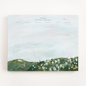 May include: A weekly planner pad with a landscape design. The top of the pad has the text "The week of GEORGIA BROOKS" and the days of the week. The bottom of the pad has a green hill with white and yellow flowers.