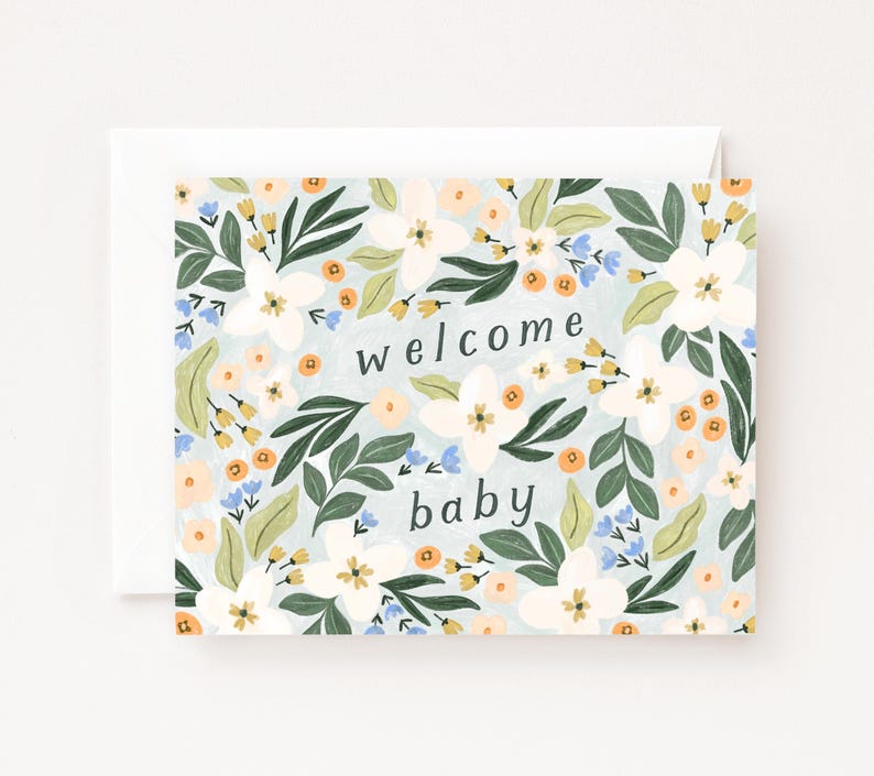Op de afbeelding: Een wenskaart met een bloemenontwerp en de woorden "welcome baby". De kaart heeft witte bloemen, groene bladeren en kleine oranje en blauwe accenten op een lichtblauwe achtergrond. De kaart zit in een witte envelop.