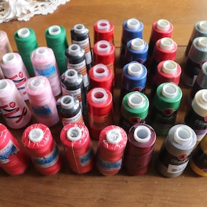 May include: A collection of colorful spools of thread, some with white labels and some with black labels. The spools are arranged in rows on a wooden surface.
