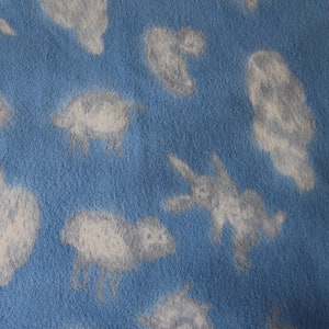 May include: A blue fleece fabric with a pattern of white sheep and cloud shapes. The sheep are depicted in a simple, cartoonish style, and the clouds are fluffy and irregular. The fabric has a soft, textured appearance.