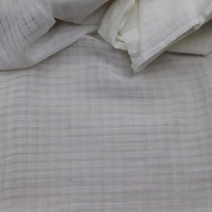 May include: A white fabric with a subtle, semi-sheer texture featuring a faint, grid-like pattern of thin, light gray horizontal and vertical lines woven into the material, creating a soft, layered appearance with visible folds and drapes.