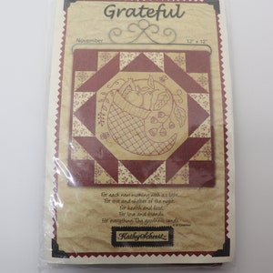 May include: A packaged quilt kit with a central design of a basket filled with fruit and leaves, surrounded by a red and tan geometric pattern. The text "Grateful" and "November" are visible, along with a quote and the designer's name.