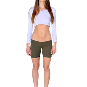 May include: A white long-sleeve crop top and olive green biker shorts. The top has a crew neck and the shorts are form-fitting. The person is wearing glasses and purple flip-flops.
