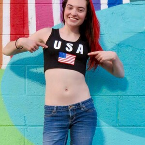 Puede incluir: Una mujer con un top negro corto con la palabra "USA" y una bandera estadounidense. Lleva pantalones vaqueros azules y tiene el pelo rojo.