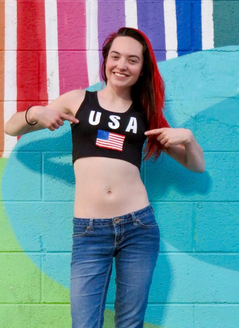 Puede incluir: Una mujer con un top negro corto con la palabra "USA" y una bandera estadounidense. Lleva pantalones vaqueros azules y tiene el pelo rojo.