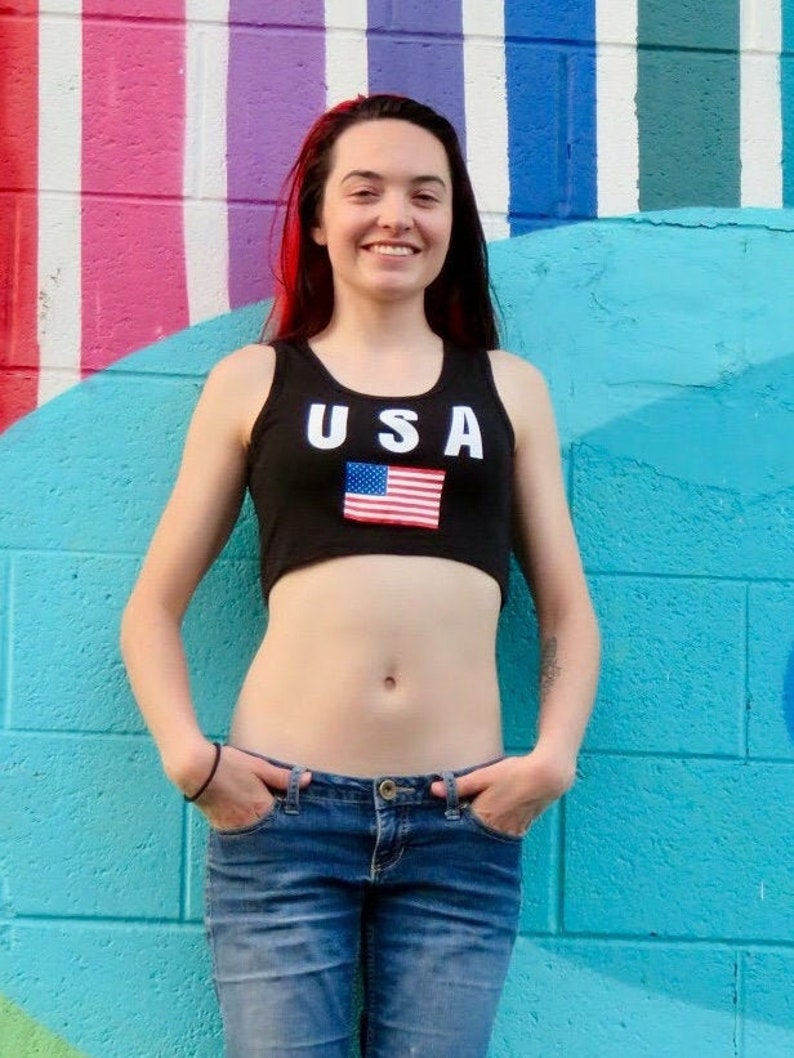 Puede incluir: Una mujer con un top negro de tirantes con la palabra "USA" y una bandera estadounidense impresa. Lleva pantalones vaqueros azules y tiene el pelo rojo.