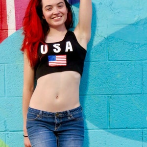 Puede incluir: Una mujer con el pelo rojo largo lleva un top corto negro con la palabra "USA" y una bandera estadounidense impresa en &eacute;l. Tambi&eacute;n lleva pantalones vaqueros azules y est&aacute; de pie frente a una pared a rayas azul y rosa.