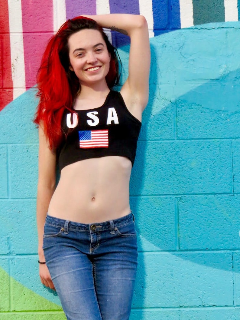 Puede incluir: Una mujer con el pelo rojo largo lleva un top corto negro con la palabra "USA" y una bandera estadounidense impresa en &eacute;l. Tambi&eacute;n lleva pantalones vaqueros azules y est&aacute; de pie frente a una pared a rayas azul y rosa.